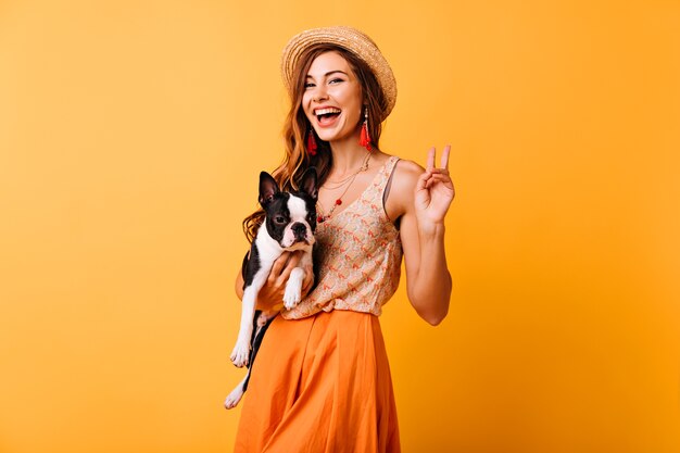 フレンチブルドッグを持って笑っているスタイリッシュな女の子。彼女の犬と一緒にスタジオでリラックスしている楽観的な赤毛の女性。