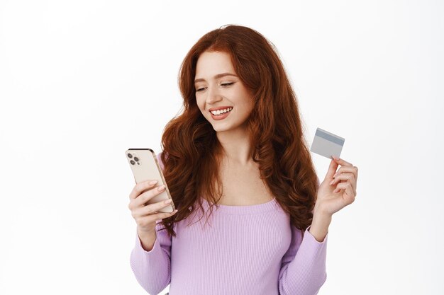 Stylish girl paying in mobile phone, order something, holding credit card with smartphone and shopping online, smiling relaxed, standing over white background