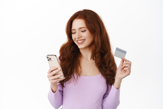 휴대전화로 결제하고, 주문하고, 스마트폰으로 신용카드를 들고 온라인 쇼핑을 하고, 편안하게 웃고, 흰색 배경 위에 서 있는 세련된 소녀