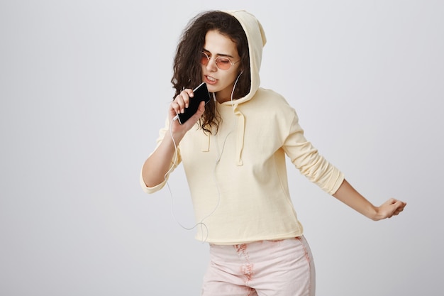 Stylish girl playing karaoke app, singing song in mobile phone, wearing earphones