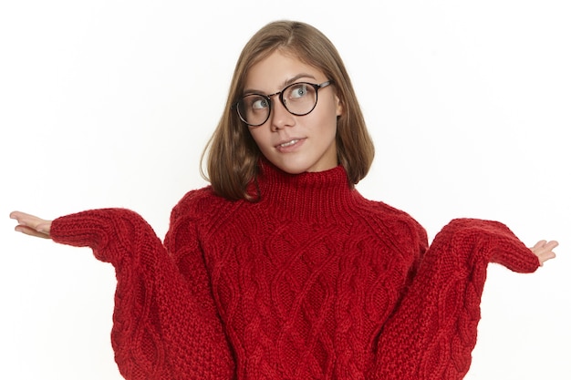 Stylish girl in spectacles and jumper shrugging shoulders, being at loss, does not know how to respond, looking up with clueless indifferent facial expression. Confused young woamn posing