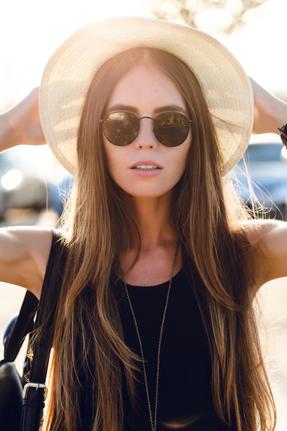 Stylish girl standing near road wearing short black dress, straw hat, black eyeglasses, and black backpack. She smiles in the warm rays of setting sun. She touches her sunglasses with hand