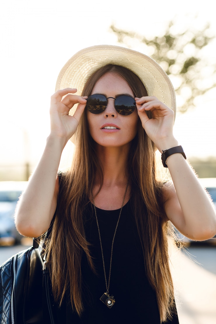 Free Photo stylish girl standing near road wearing short black dress, straw hat, black eyeglasses, and black backpack. she smiles in the warm rays of setting sun. she touches her sunglasses with hand