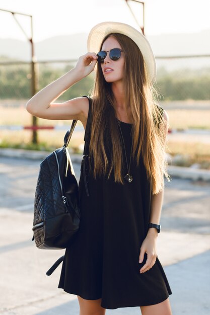 Stylish girl standing near road wearing short black dress, straw hat, black eyeglasses, and black backpack. She smiles in the warm rays of setting sun. She touches her sunglasses with hand