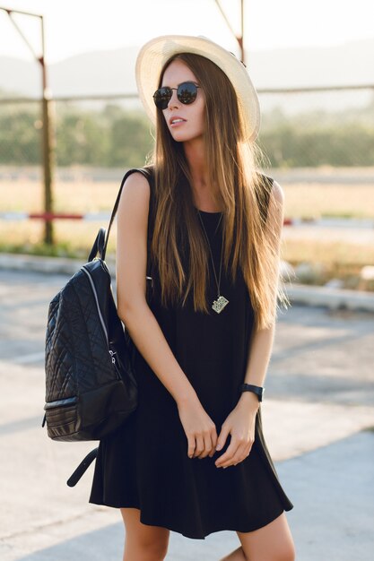 Stylish girl standing near road wearing short black dress, straw hat, black eyeglasses, and black backpack. She smiles in the warm rays of setting sun