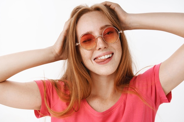 Stylish girl in trendy sunglasses and pink t-shirt holding hands spread on head winking, sticking out tongue happily and smiling from joy and relaxed emotions having fun, relaxing over gray wall