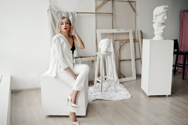 Стильная девушка в белом костюме сидит на белом кубе в галерее