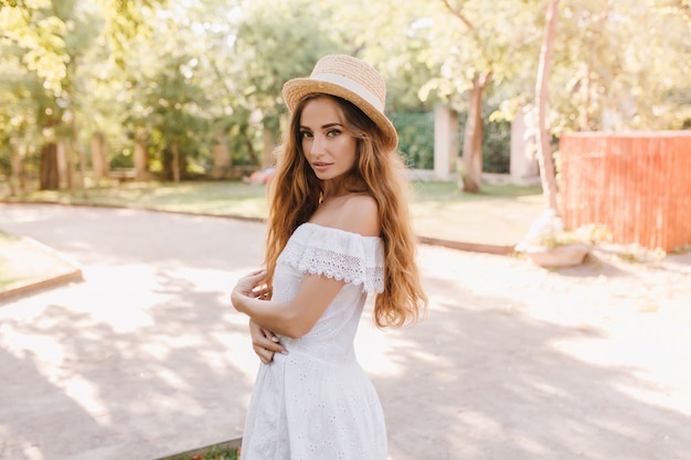 Stylish girl with blonde curls looking over shoulder during walk in park. Slim young lady in beautiful white dress enjoying sunshine in summer morning.