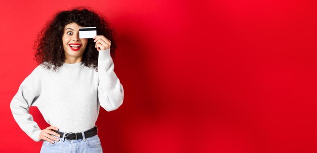 Stylish girl with curly hair showing plastic credit card over eye and smiling standing against red b