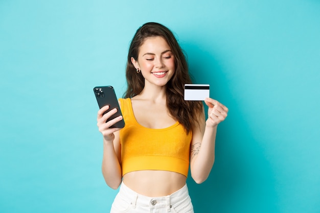 Стильная девушка с пластиковой кредитной картой улыбается, оплачивает онлайн-заказ, делает покупки в смартфоне, стоя на синем фоне. Копировать пространство