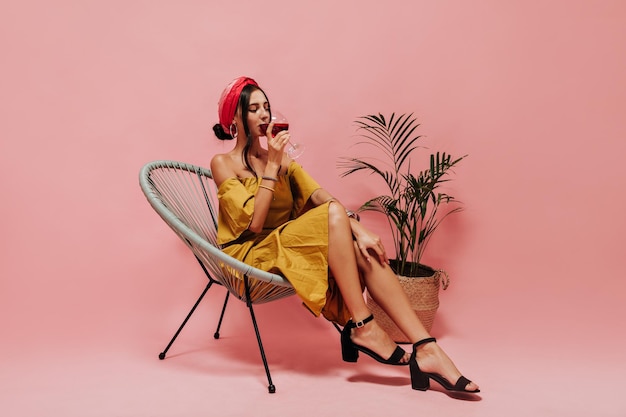 Stylish girl in yellow outfit black shoes and red bandana drinking wine with closed eyes and sitting on modern chair on pink backdrop