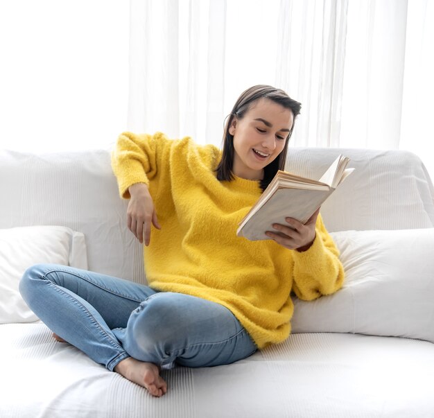 黄色いセーターを着たスタイリッシュな女の子が、本を持って自宅のソファで休んでいます。