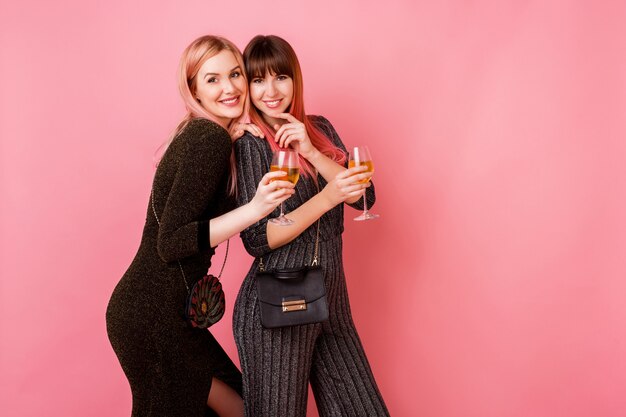 Стильные девушки в бокалах с алкогольными напитками позируют на светло-розовой стене