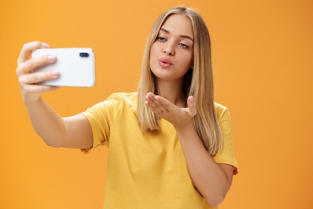 Стильная гламурная модная блогерша заканчивает запись видео через смартфон, отправляя воздушный поцелуй в камеру, делая селфи с чувственным и уверенным взглядом на экране, позируя над оранжевой стеной Premium Фотографии