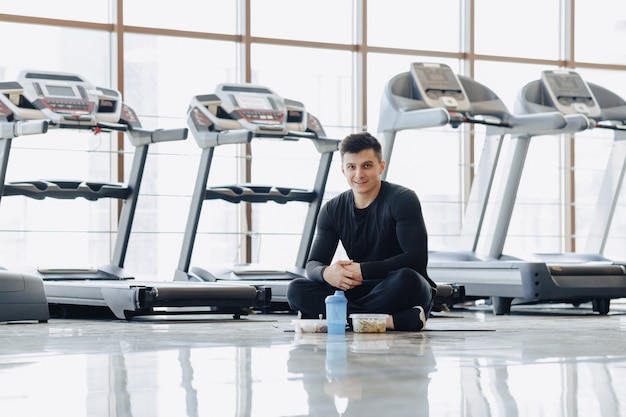 Stylish guy in the gym relaxing on the floor and eating healthy food.