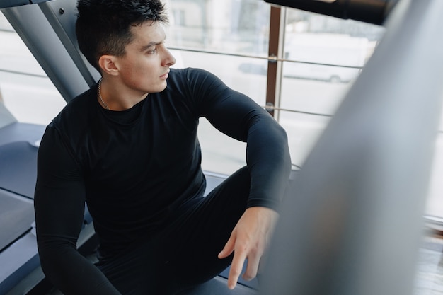 Ragazzo alla moda in palestra si siede a riposo sul tapis roulant. uno stile di vita sano.