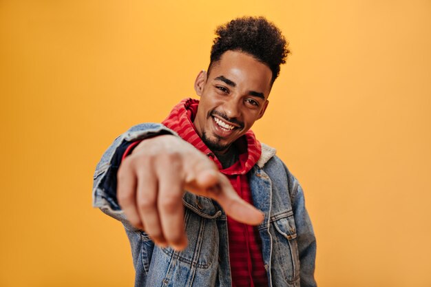 Stylish guy in red sweatshirt points his finger at camera and posing on isolated wall