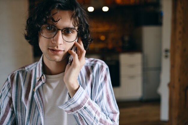 Stylish guy with curly hair posing against cozy kitchen interior having phone conversation using mobile