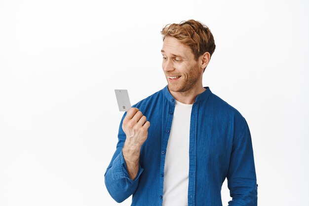 Stylish guy with red hair looks satisfied at credit card in hands smiling pleased shopping or paying contactless standing over white background