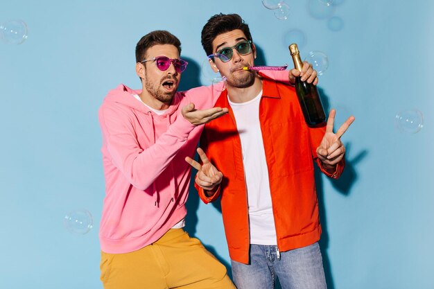 Stylish guys in colorful outfits celebrate and have fun on blue background Man in pink hoodie holds champagne bottle Guy in orange jacket blows into party horn