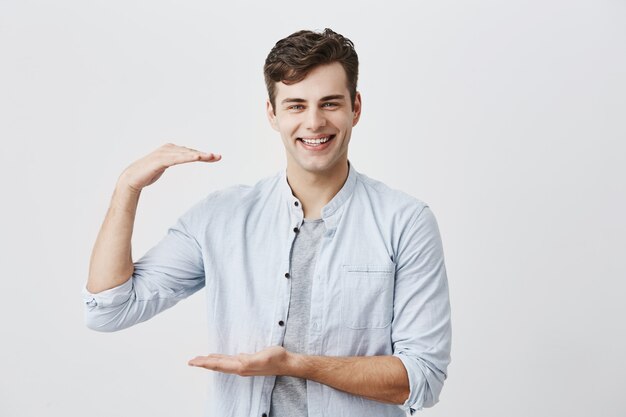 Stylish handsome guy wearing blue shirt over T-shirt showing size of something small with hands, gesturing, broadly smiling with white teeth. Dark-haired male posing 