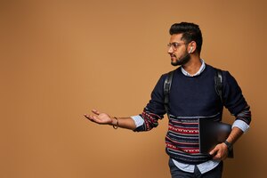 Stylish handsome indian student with laptop and backpack pointiong his hand to wall.