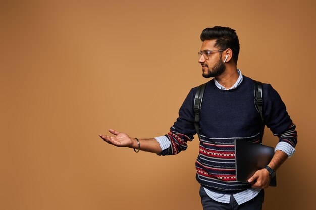 Стильный красивый индийский студент с компьтер-книжкой и рюкзаком pointiong его рука к стене.