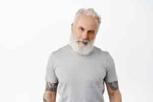 Free photo stylish handsome mature man with tattooed body and long beard, tilt head and look confident macho at front, staring serious, standing in grey t-shirt against white wall