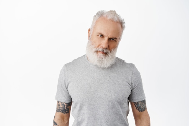 Stylish handsome mature man with tattooed body and long beard, tilt head and look confident macho at front, staring serious, standing in grey t-shirt against white wall