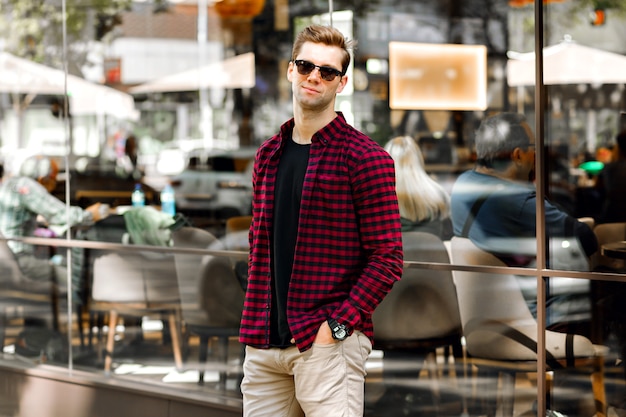 Stylish handsome young businessman sitting on the street, amazing smile, brown hairs and eyes, wearing hipster plaid shirt and beige trousers, sunglasses and watches, posing near restaurant.