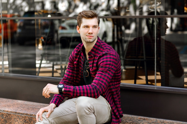 Stylish handsome young businessman sitting on the street, amazing smile, brown hairs and eyes, wearing hipster plaid shirt and beige trousers, sunglasses and watches, posing near restaurant.