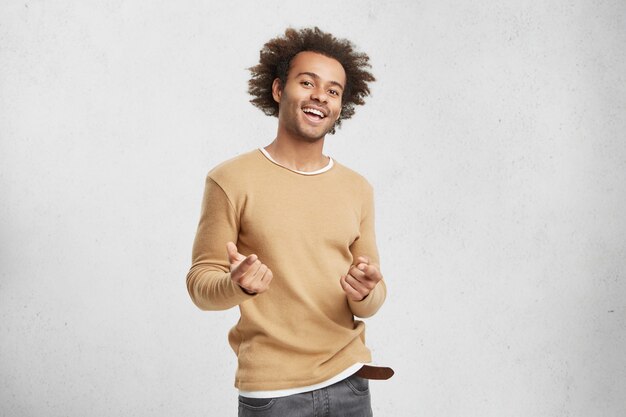 スタイリッシュな幸せなアフリカ系アメリカ人男性がカジュアルな服を着て、人差し指でカメラのポイント