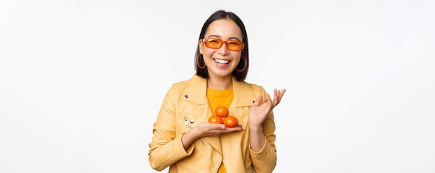Стильная счастливая азиатская девушка в солнцезащитных очках, держащая мандарины и улыбающаяся, позирующая на белом фоне