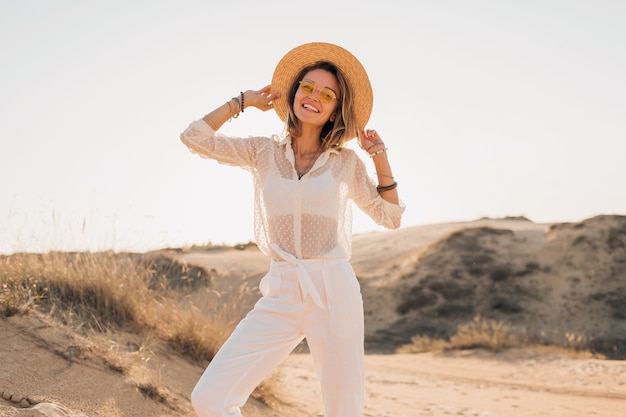 Elegante donna sorridente attraente felice in posa nella sabbia del deserto vestita in abiti bianchi che indossa cappello di paglia e occhiali da sole sul tramonto, soleggiata giornata estiva