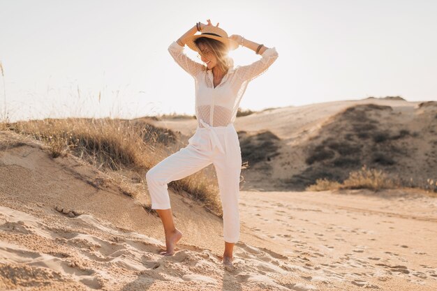 Стильная счастливая привлекательная улыбающаяся женщина позирует в песке пустыни, одетая в белую одежду, в соломенной шляпе и солнцезащитных очках на закате