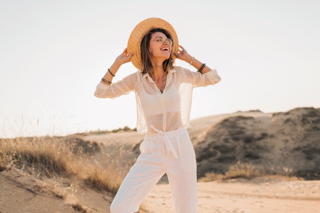 Stylish happy beautiful smiling woman posing in desert sand in white outfit wearing straw hat and sunglasses on sunset