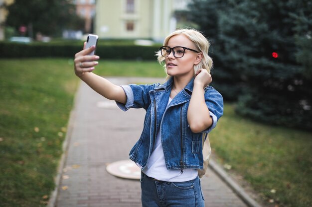Stylish happy pretty fashion blonde girl woman in jeans suite makes selfie on her phone in the city in the morning