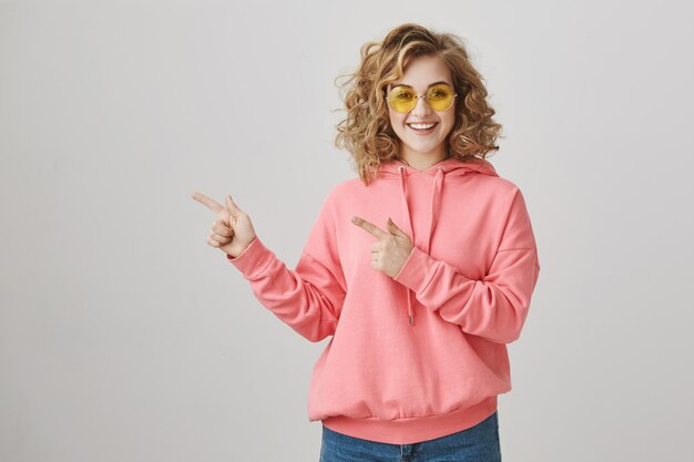 Stylish happy teenage girl in sunglasses showing way, promote advertisement