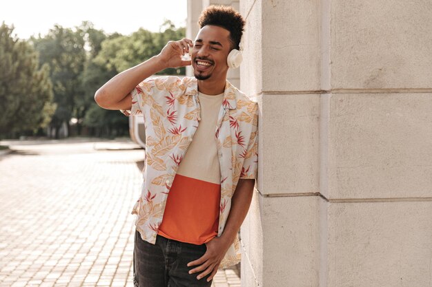 Stylish happy young man in colorful tshirt leans on wall outside Cool guy in floral shirt listens to music in headphones outdoors