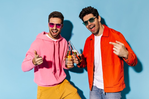 Stylish happy young men in colorful sunglasses rejoice on blue background Friends in bright outfits clink beer bottles