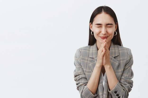 Stylish happy young woman with dark hair in jacket, closing eyes and smiling joyfully while holding hands in pray and hoping for better