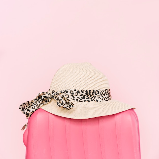 Stylish hat on plastic luggage travel bag against pink background