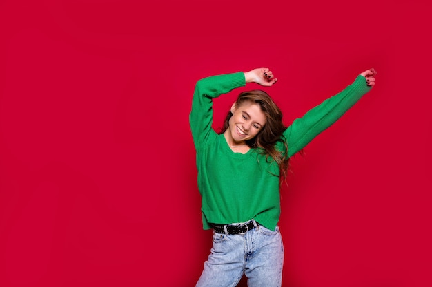 Stylish hippie girl on red pulling up hands, celebrating new year, wearing green pullover and jeans, happy carnival disco party, sparkling confetti, holding glass, having fun