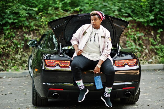 Stylish hipster arab man guy in hat posed outdoor in street and sitting on the trunk his black muscle car