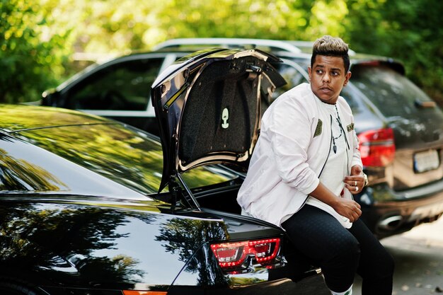 Stylish hipster arab man guy posed outdoor in street and sitting on the trunk his black muscle car Rich black man