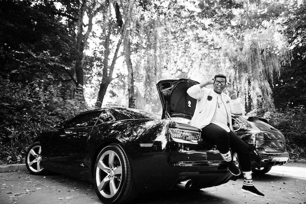 Стильный хипстер-араб в солнцезащитных очках позирует на улице и сидит на багажнике своего черного мускулистого автомобиля Богатый темнокожий мужчина