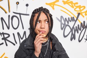 Stylish hipster girl has dreadlocks being deep in thoughts keeps hand near mouth concentrated above poses in urban environment against graffiti wall wears black jacket