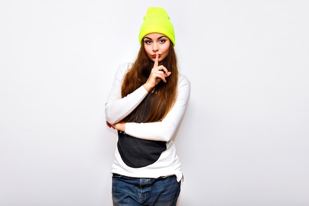 Donna alla moda hipster in posa contro il muro bianco, orario invernale, maglione, cappello al neon e jeans, abbigliamento sportivo alla moda casual, capelli lunghi, trucco luminoso, flash, viso sexy serio.