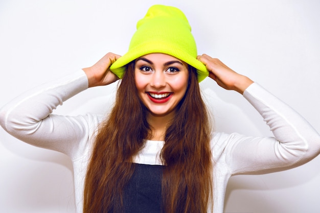 Free photo stylish hipster woman posing against white wall, winter time, sweater, neon hat and jeans, casual trendy sportive outfit, long hairs, bright make up, flash, serious sexy face.