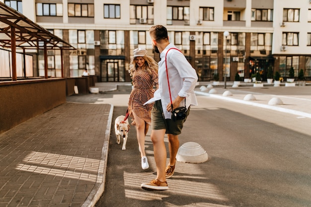 Stylish husband and wife in summer outfits run and play with their dog on background of apartment house. Man in light shirt holds his beloved hand and carries camera.